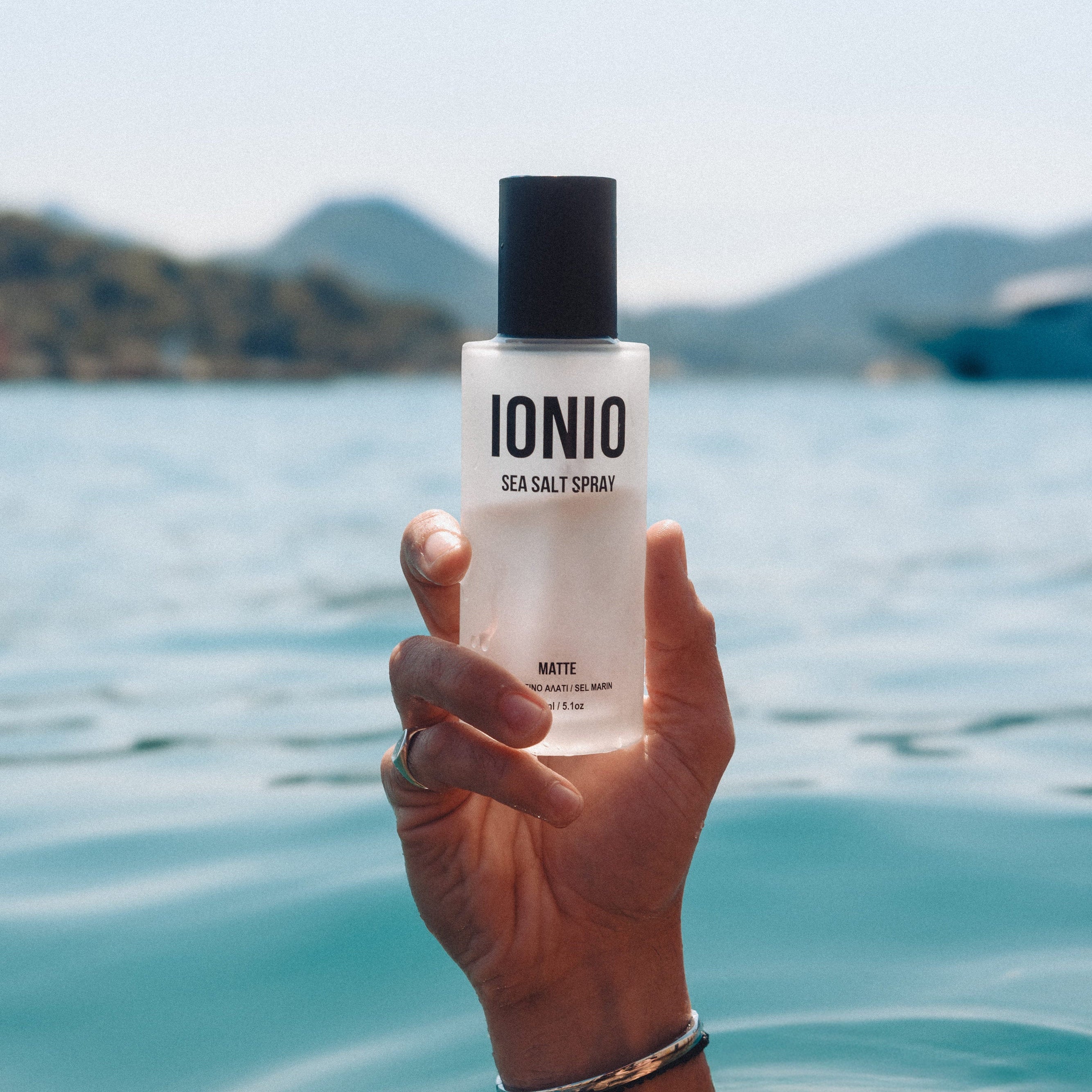Hand holding a bottle of 'IONIO' sea salt spray over water with mountains in the background